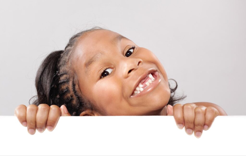 A very happy young girl peeking up from behind a white surface, smiling with a couple of missing teeth.