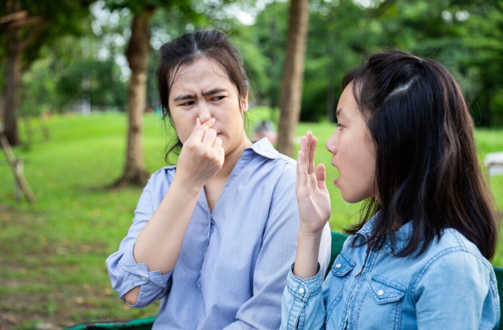 A mom wrinkles her nose in disapproval at her daughter's bad breath, while the daughter exhales onto her hand to catch a whiff herself.